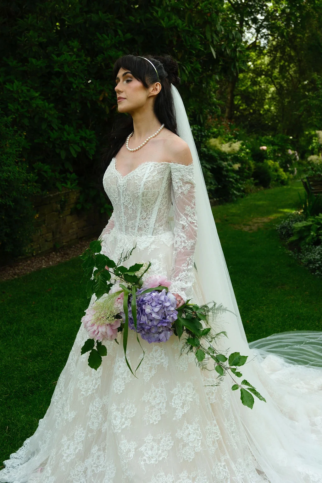A bride in an off-the-shoulder lace gown with long sleeves poses in a lush garden, holding a cascading bouquet of purple hydrangeas and pink flowers.