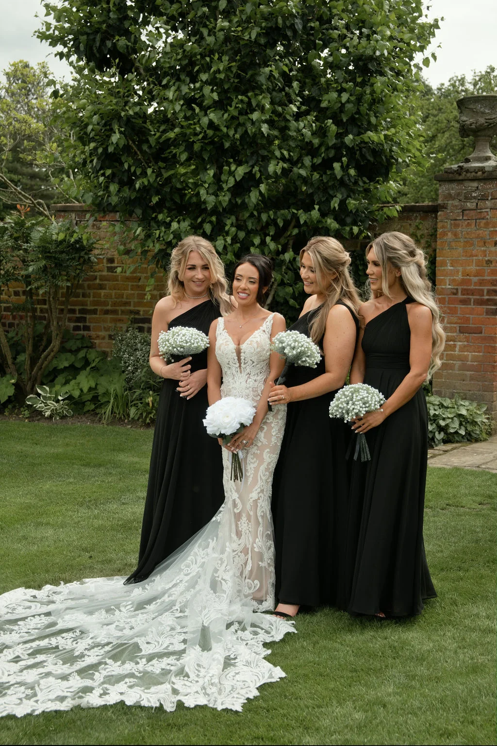 A stunning bride in an intricate lace gown with a dramatic long train poses outdoors on a lawn with her four bridesmaids wearing elegant black one-shoulder dresses and holding baby's breath bouquets.