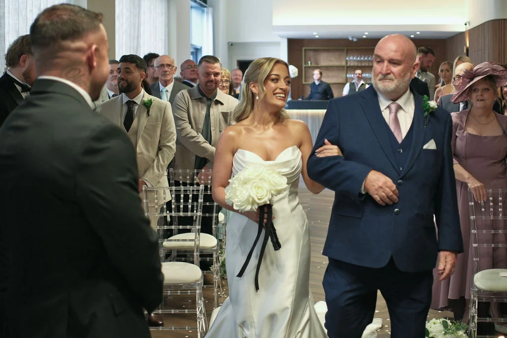 A smiling bride walks down the aisle arm in arm with her father towards the waiting groom, holding a bouquet of white roses.