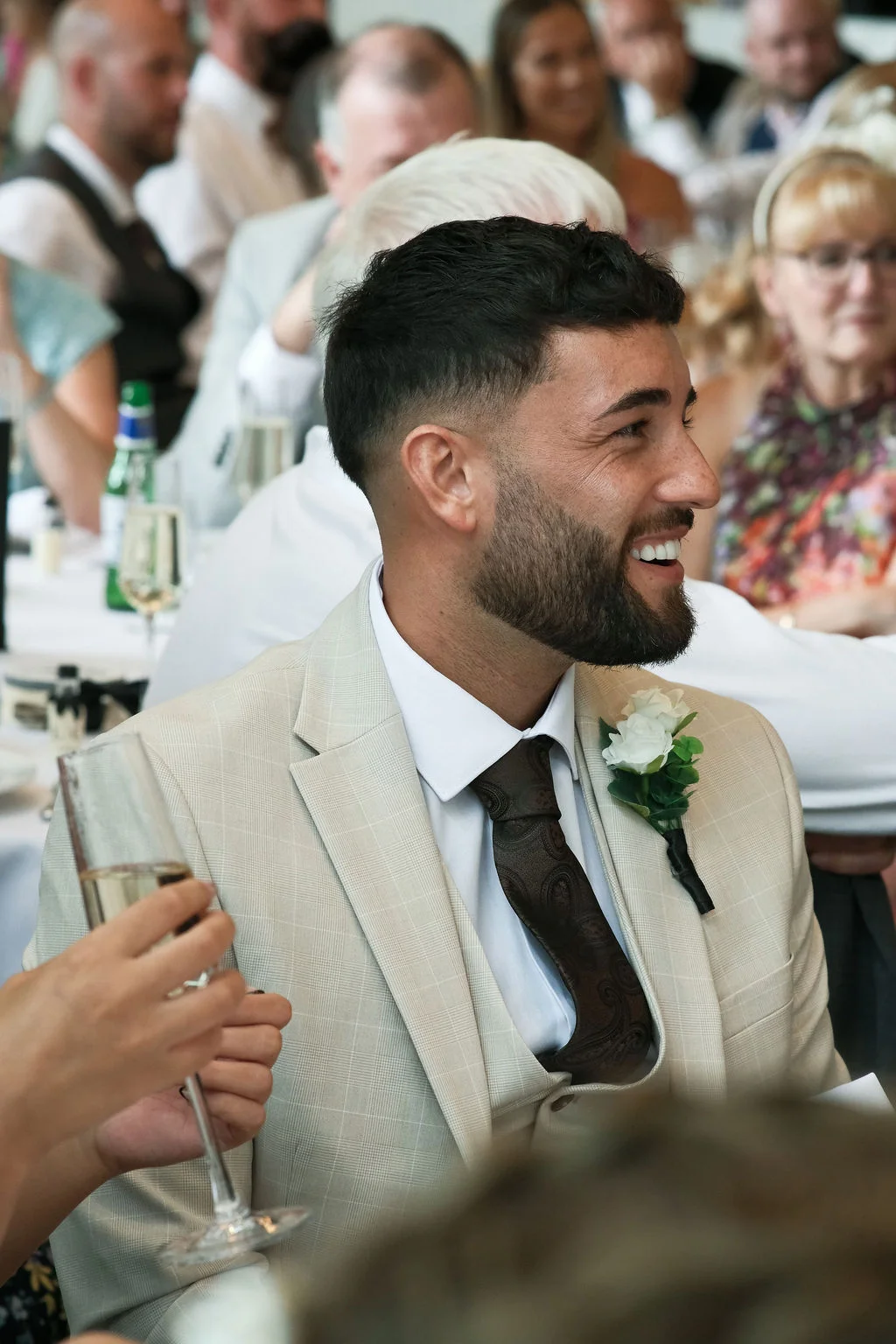A smiling groom in a beige checked three-piece suit with a patterned tie and white buttonhole laughs during wedding speeches while holding a glass of champagne.