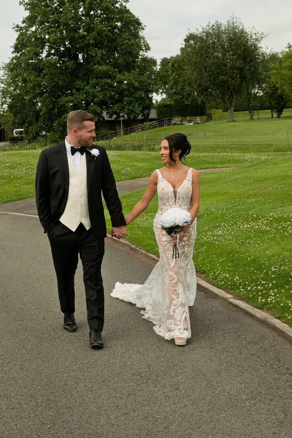 A newlywed couple holds hands and smiles at each other while walking along a paved path bordered by lush green lawns.