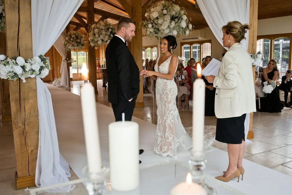 A bride and groom exchange rings during a civil ceremony inside a light-filled oak framed barn adorned with white florals and draping.