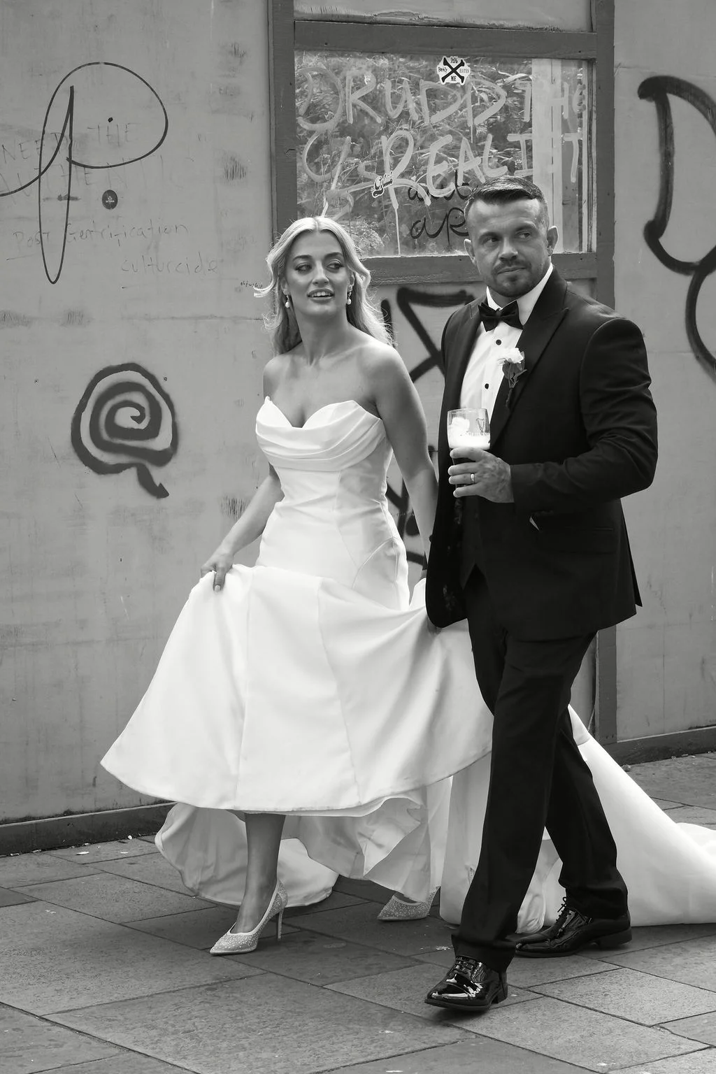 A stylish newlywed couple walks down a city street past a graffiti-covered wall, with the bride holding up her white satin gown and the groom holding a drink.