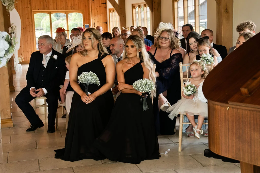 Two bridesmaids in matching black one-shoulder dresses holding posy bouquets sit attentively among wedding guests inside a rustic oak-framed barn.