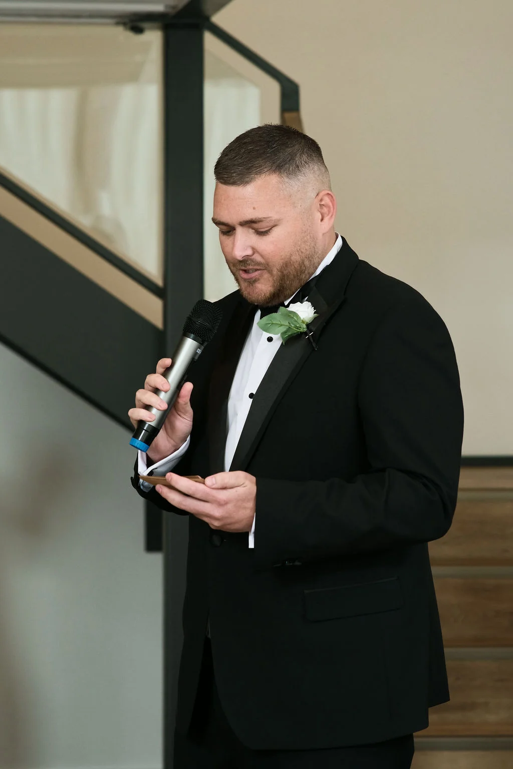 A groom in a black tuxedo delivers a heartfelt wedding speech while holding a microphone and notes indoors.