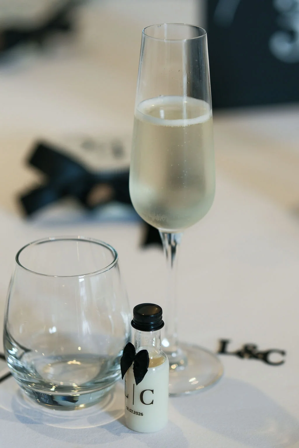 A close-up view of a wedding table setting featuring a champagne flute, a water glass, and a miniature favour bottle adorned with a black bow.