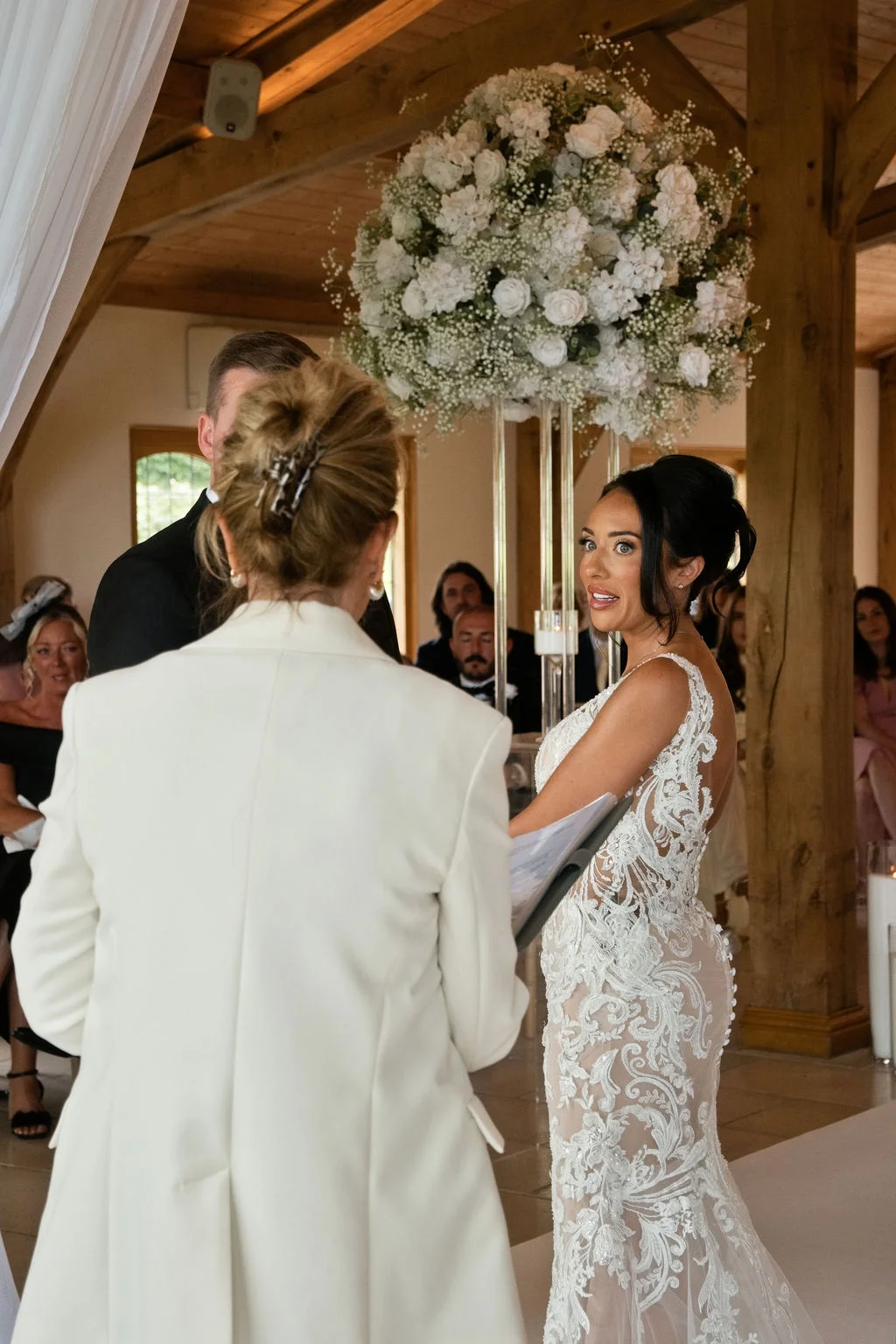 A bride in a sheer lace gown looks back toward her guests with a warm expression during a civil ceremony inside a converted oak-framed barn.