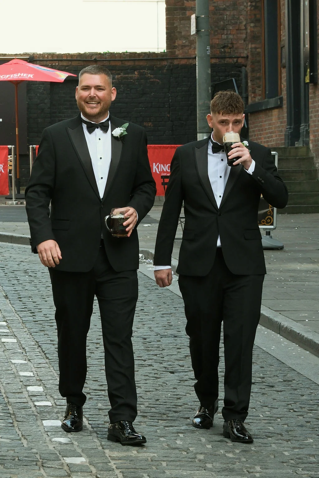 Two groomsmen in black tuxedos and bow ties stroll down a cobbled city street while enjoying pints of dark beer.