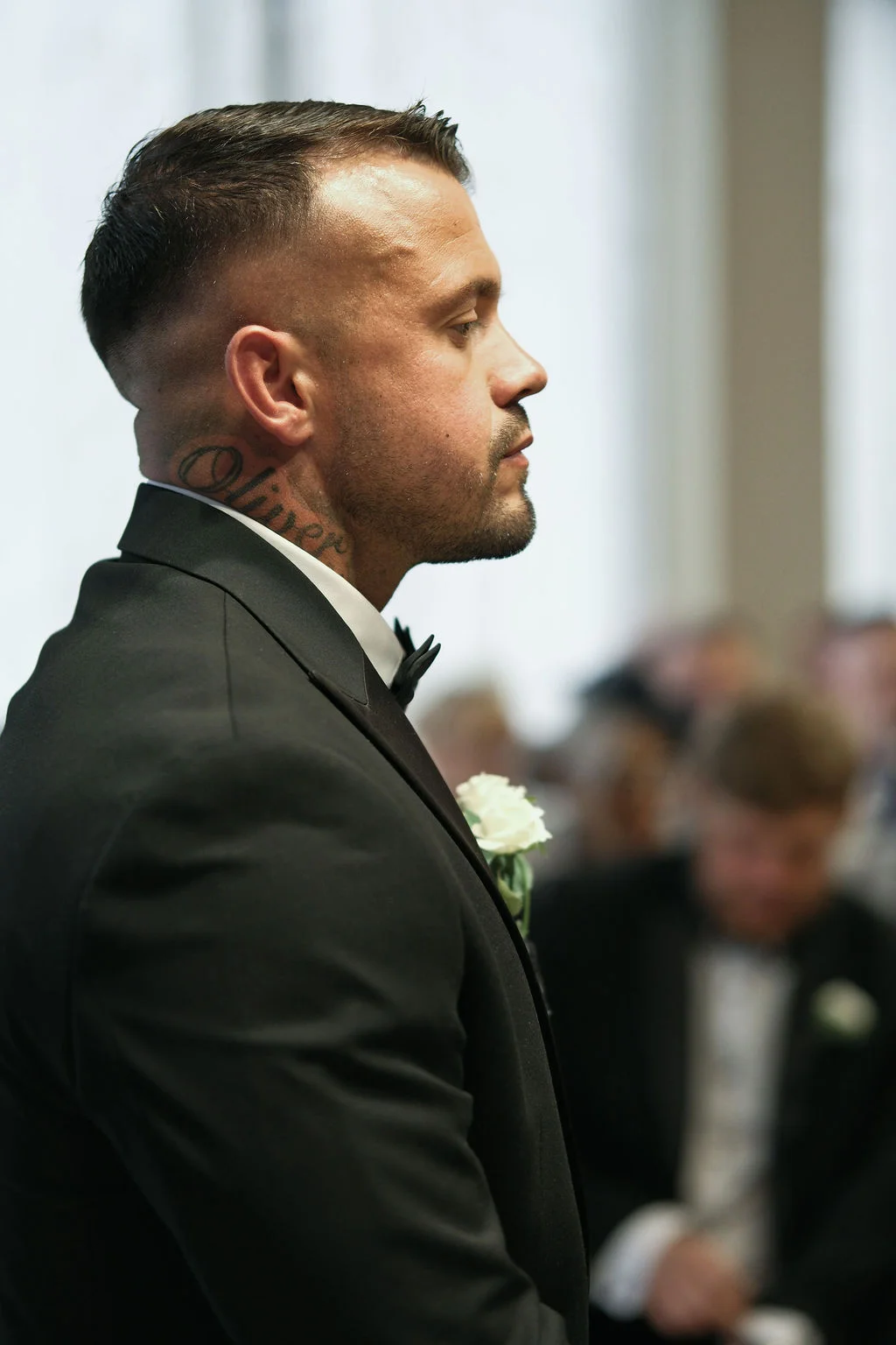A groom stands at the altar in a black tuxedo with a white boutonniere, captured in profile with a shallow depth of field during the ceremony.