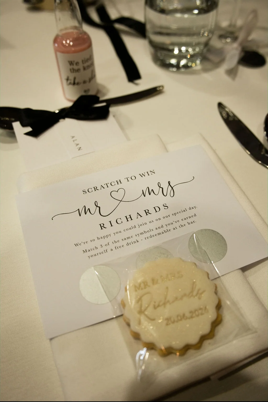 A detailed place setting features a custom scratch card favour and an iced biscuit resting on a crisp white napkin.