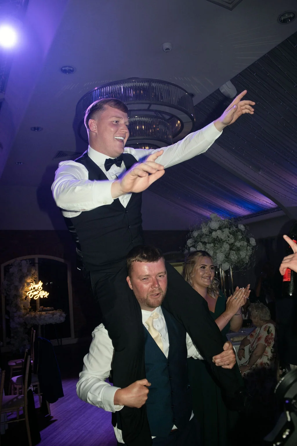 A joyous wedding guest is lifted onto another man's shoulders on a lively dance floor beneath a lit chandelier.