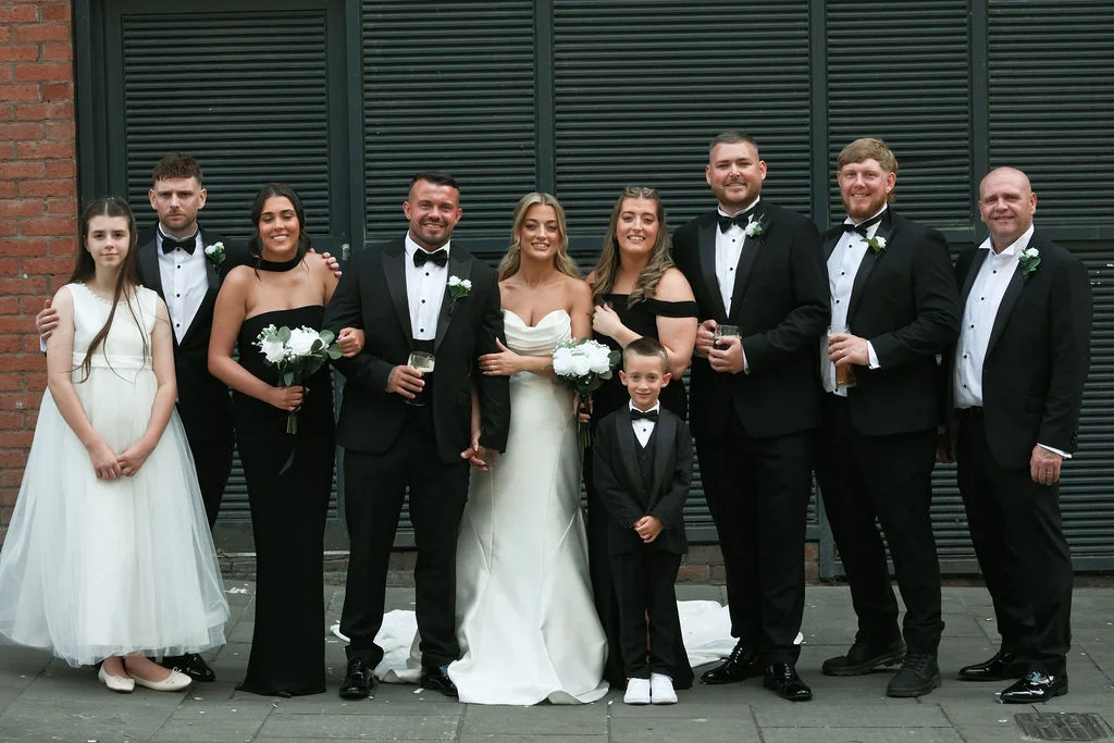 A newlywed couple poses with their wedding party of groomsmen in black tuxedos, bridesmaids in black gowns, and a flower girl and page boy in front of a dark slatted background.