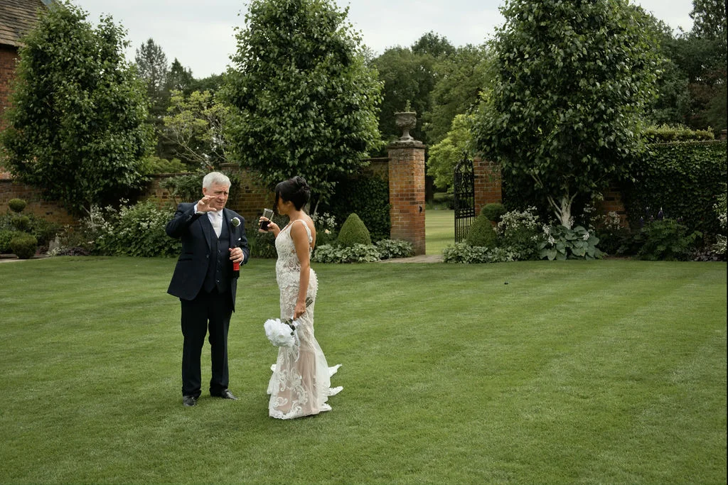 A bride in a lace gown holding a bouquet stands on a manicured lawn having a conversation with her father outside a country house venue.