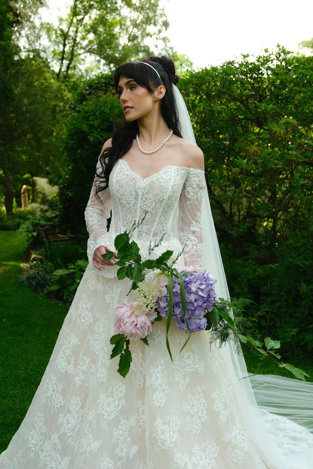 A bride stands in a lush garden holding a cascading bouquet of purple hydrangeas and soft pink flowers, wearing an off-the-shoulder lace a-line gown and a pearl necklace.