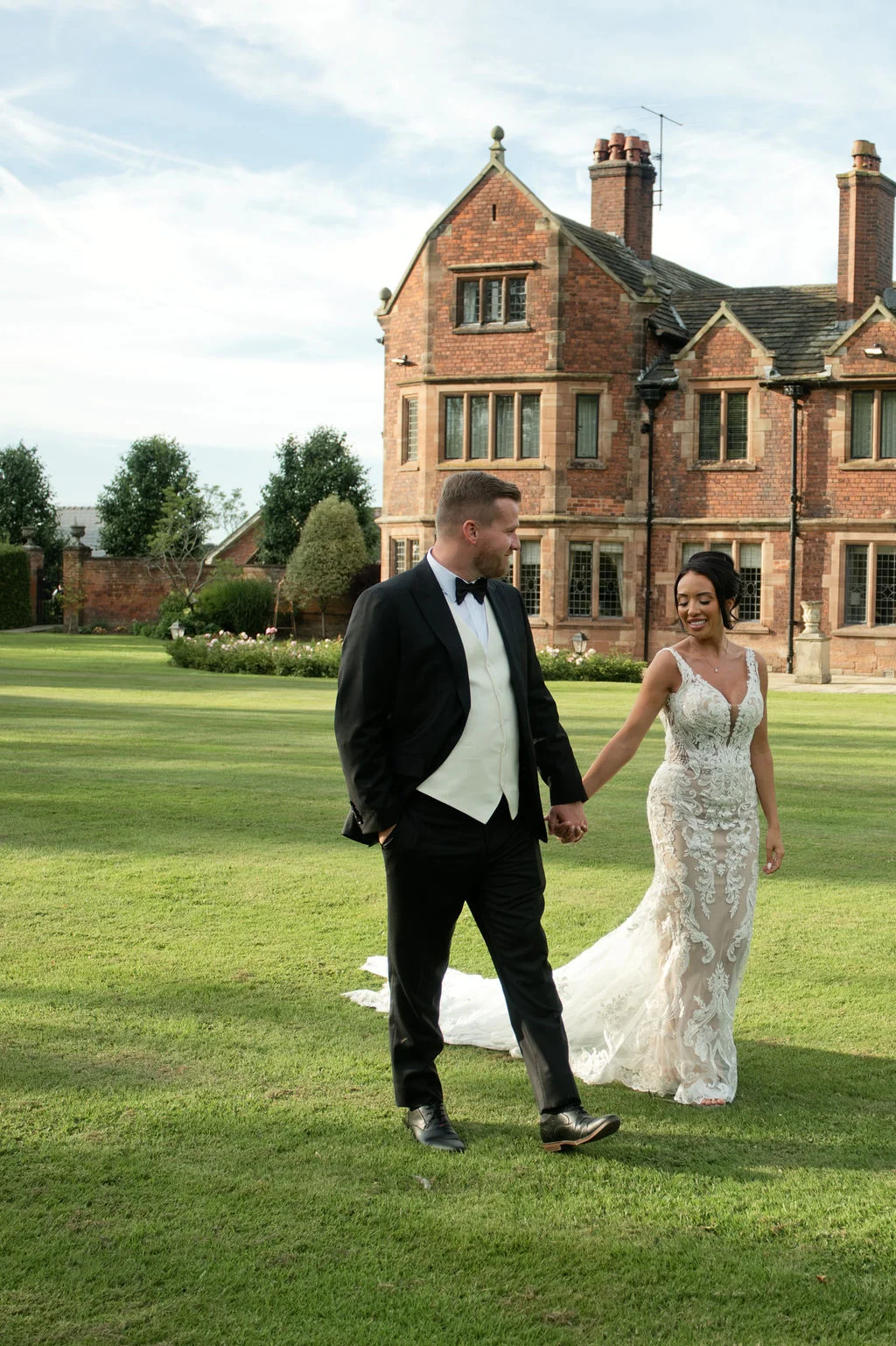 A bride and groom walk hand in hand across the manicured lawn of a grand red brick manor house on their wedding day.