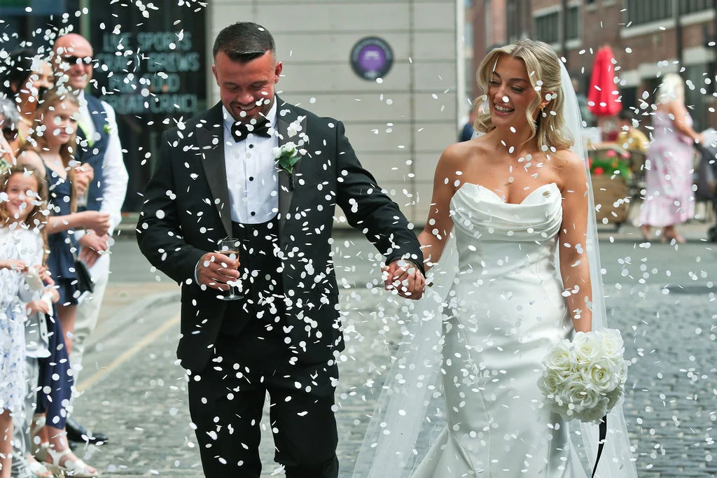 A joyful newlywed couple walks hand in hand through a city street amidst a shower of white confetti, with the groom in a classic black tuxedo and the bride in an elegant strapless satin gown holding a white rose bouquet.