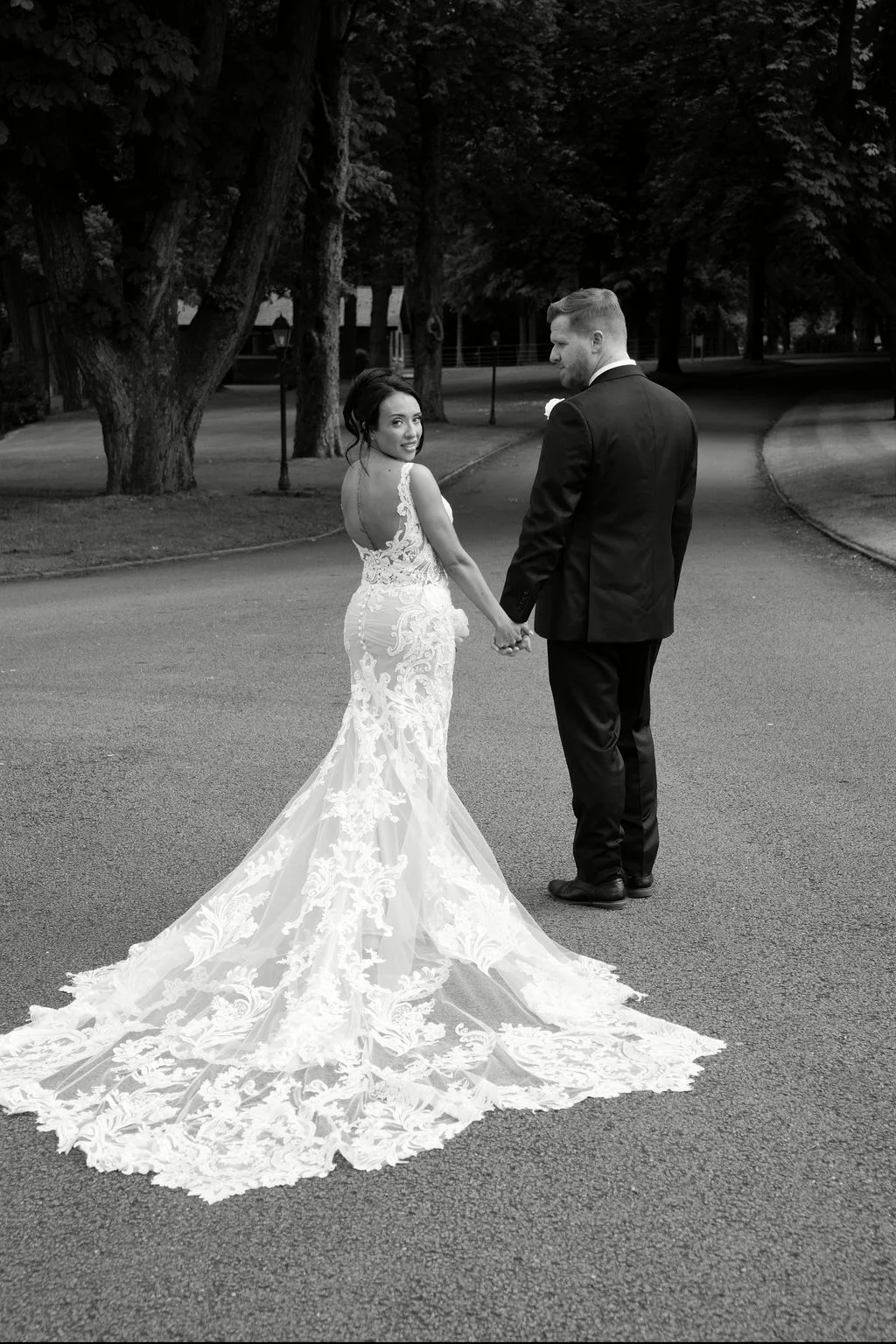 Newlyweds walking hand in hand along a tree-lined pathway, with the bride glancing back over her shoulder wearing a dramatic lace gown with a long train.