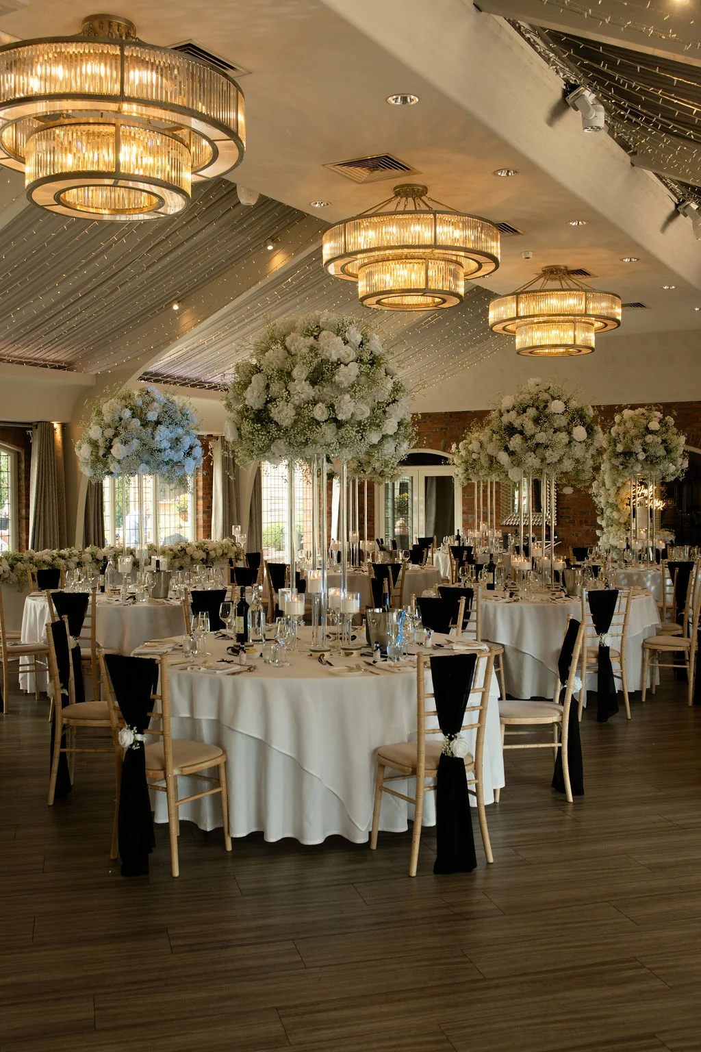 A grand hotel ballroom reception featuring round tables draped in white linens, elegant tall floral centerpieces with white roses and baby's breath, and dramatic crystal chandeliers illuminating the ceiling canopy.
