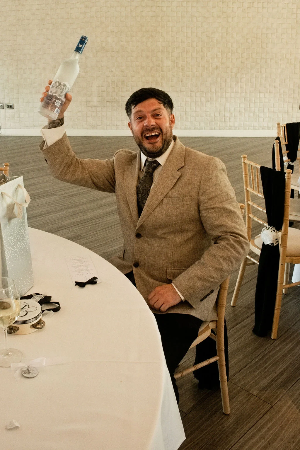 A joyful groom wearing a tan suit and patterned tie sits at a white-clothed reception table, excitedly raising a bottle of vodka in the air while laughing heartily.