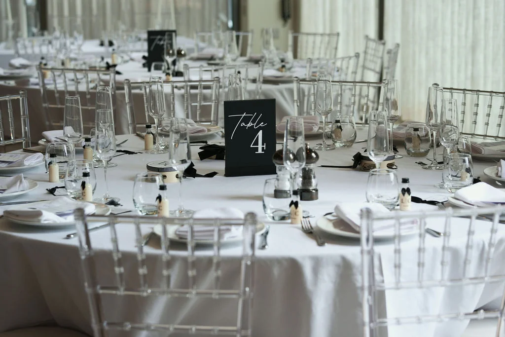A sophisticated monochrome wedding reception tablescape features crisp white linens, clear ghost chairs, and sleek black table numbers.