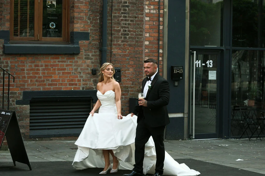 A newlywed couple walks together outside an urban venue with a red brick exterior, the bride lifting her high-low satin wedding dress while the groom carries a drink in his black tuxedo.