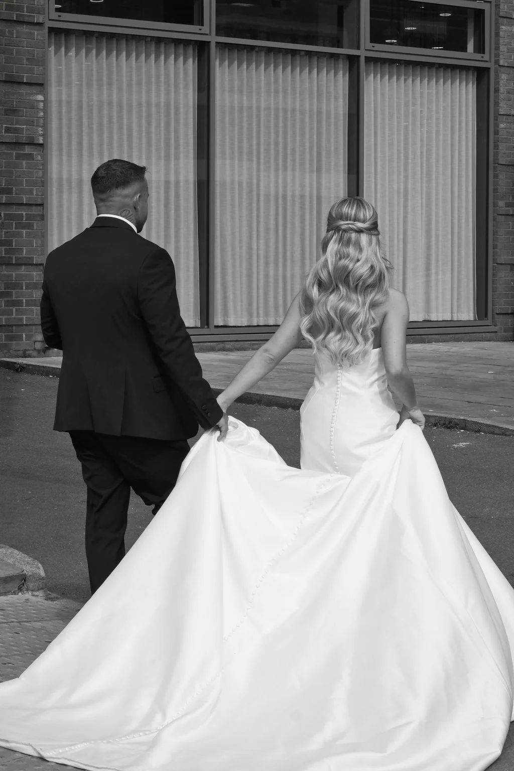 Newlyweds seen from behind walking away while holding hands outside a modern brick building, with the bride displaying her flowing train and half-up wavy hairstyle.