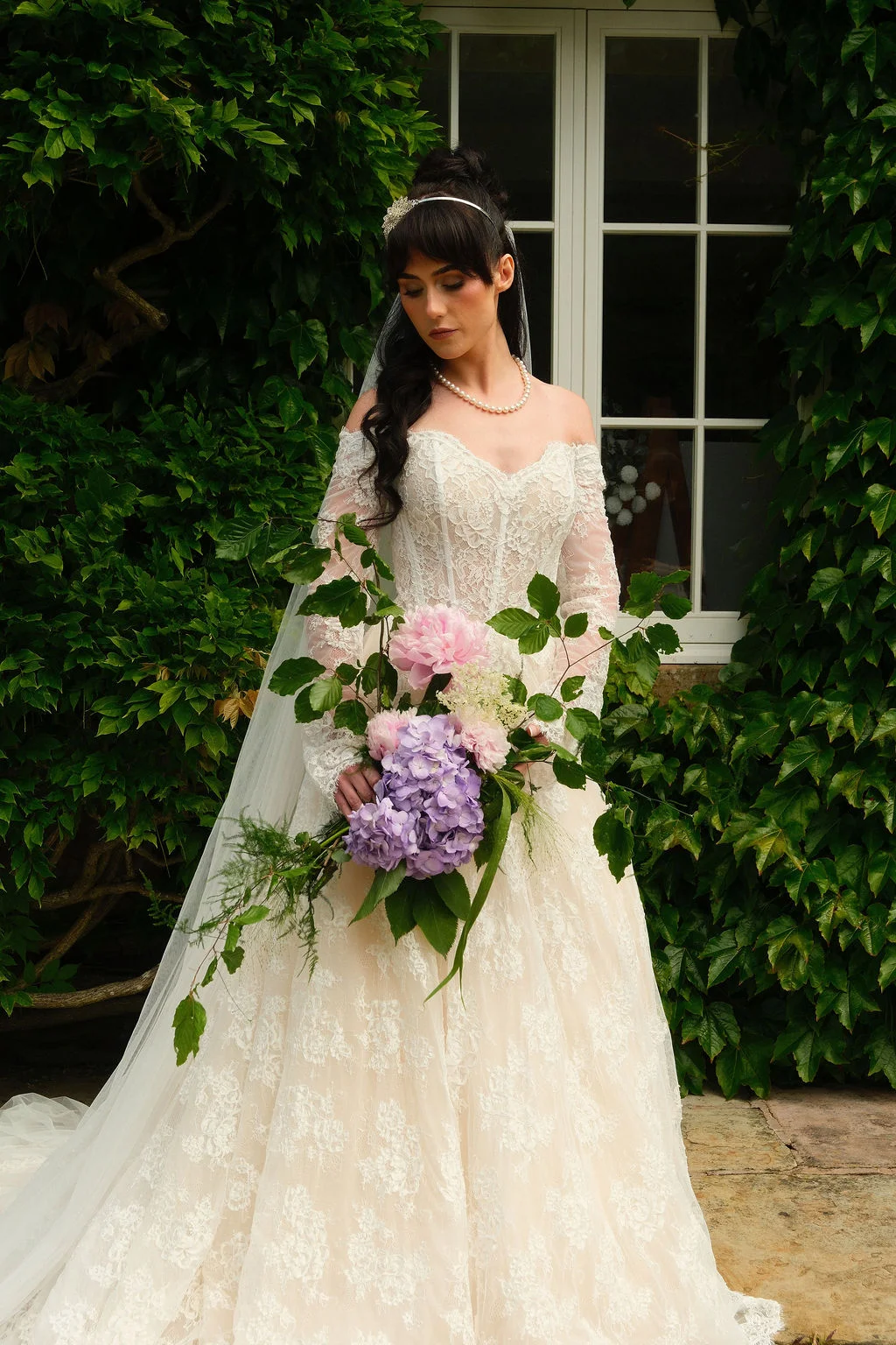 A bride in an off-the-shoulder lace a-line gown stands before an ivy-covered wall, holding a cascading bouquet of pink peonies and purple hydrangeas.