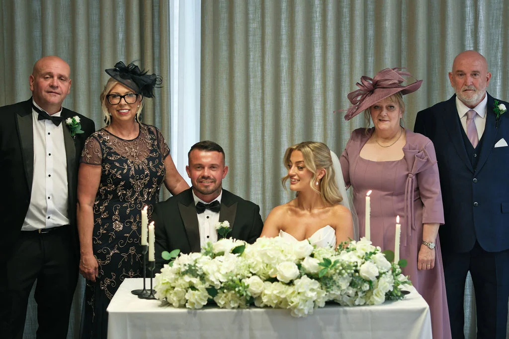 The newly married couple sits together at a white-clothed table adorned with lush white floral arrangements and candles, flanked by family members in formal wedding attire.