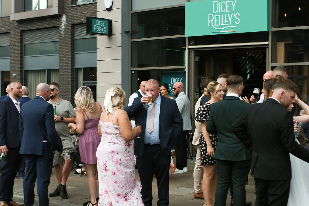 Wedding guests mingle outside a modern pub venue during the daytime drinks reception, chatting and holding drinks.