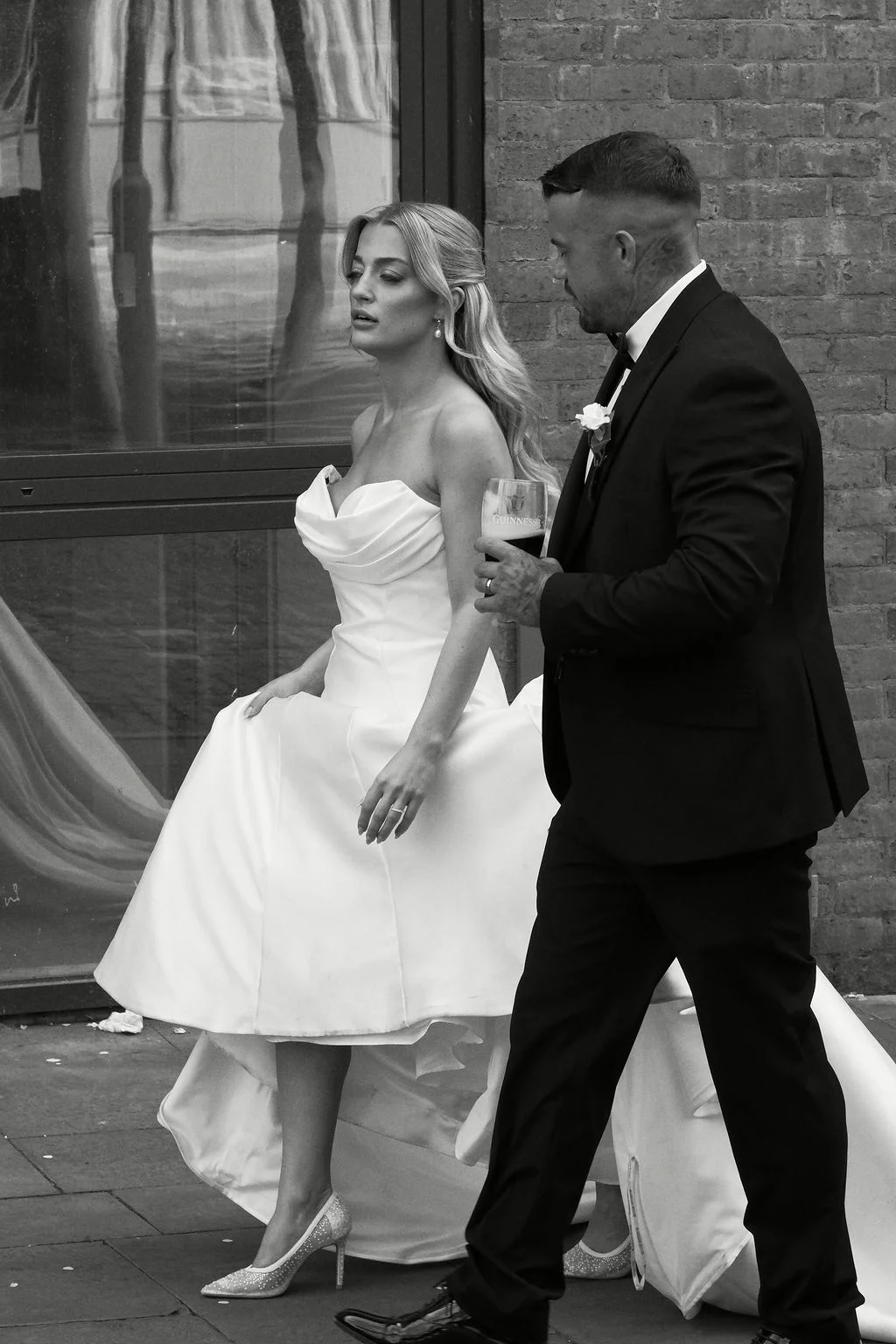 A bride in a strapless high-low wedding dress and sparkly heels walks alongside a groom in a black tuxedo holding a drink.