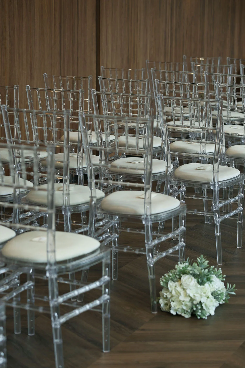 An empty wedding ceremony space featuring rows of clear ghost chairs and a bouquet of white roses and greenery on the wooden floor.
