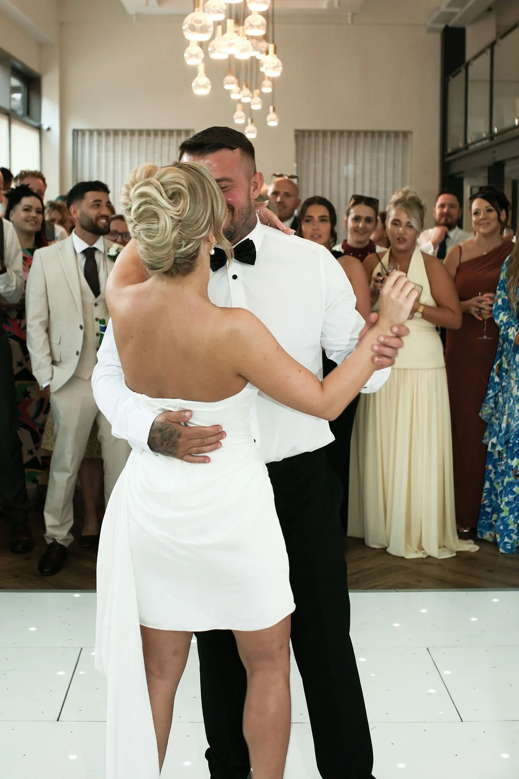 Newlyweds share their first dance on a glowing white dance floor while surrounded by smiling wedding guests.
