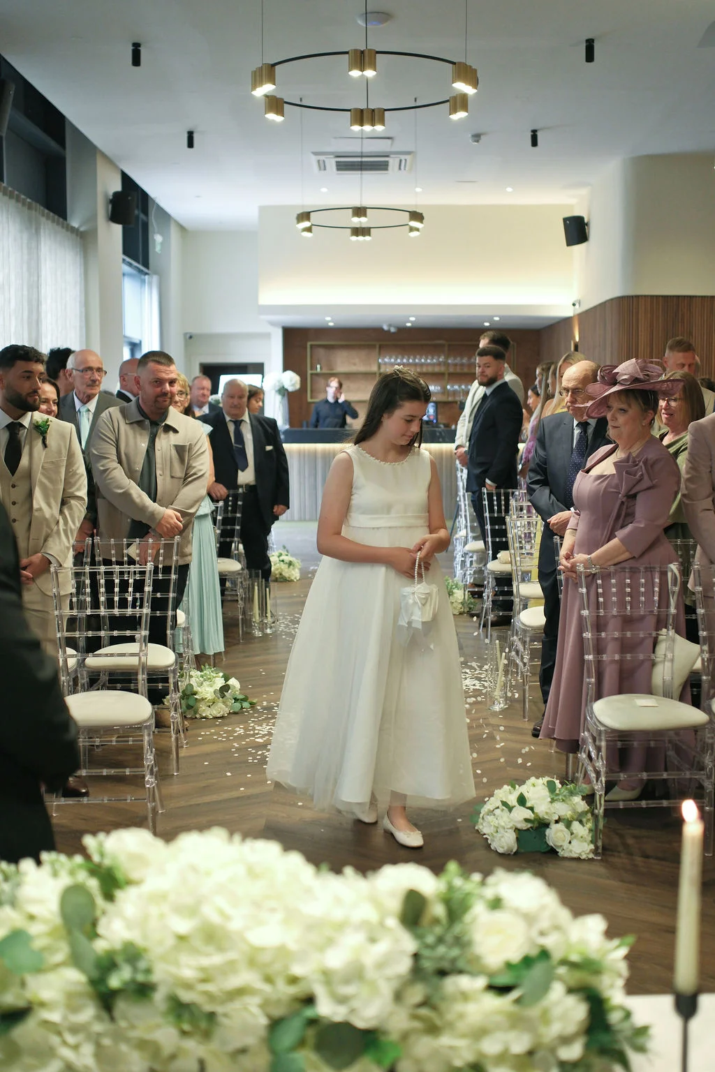 A young flower girl in a white dress walks down the aisle during an indoor wedding ceremony while guests look on.