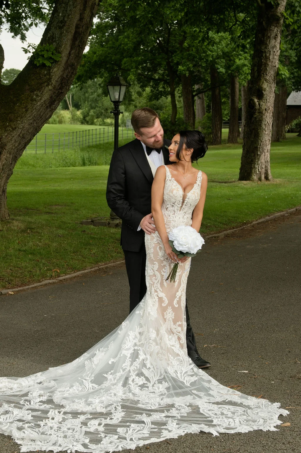 A bride in a lace mermaid wedding dress with a dramatic train and a groom in a black tuxedo pose lovingly on a driveway surrounded by trees and green lawns.