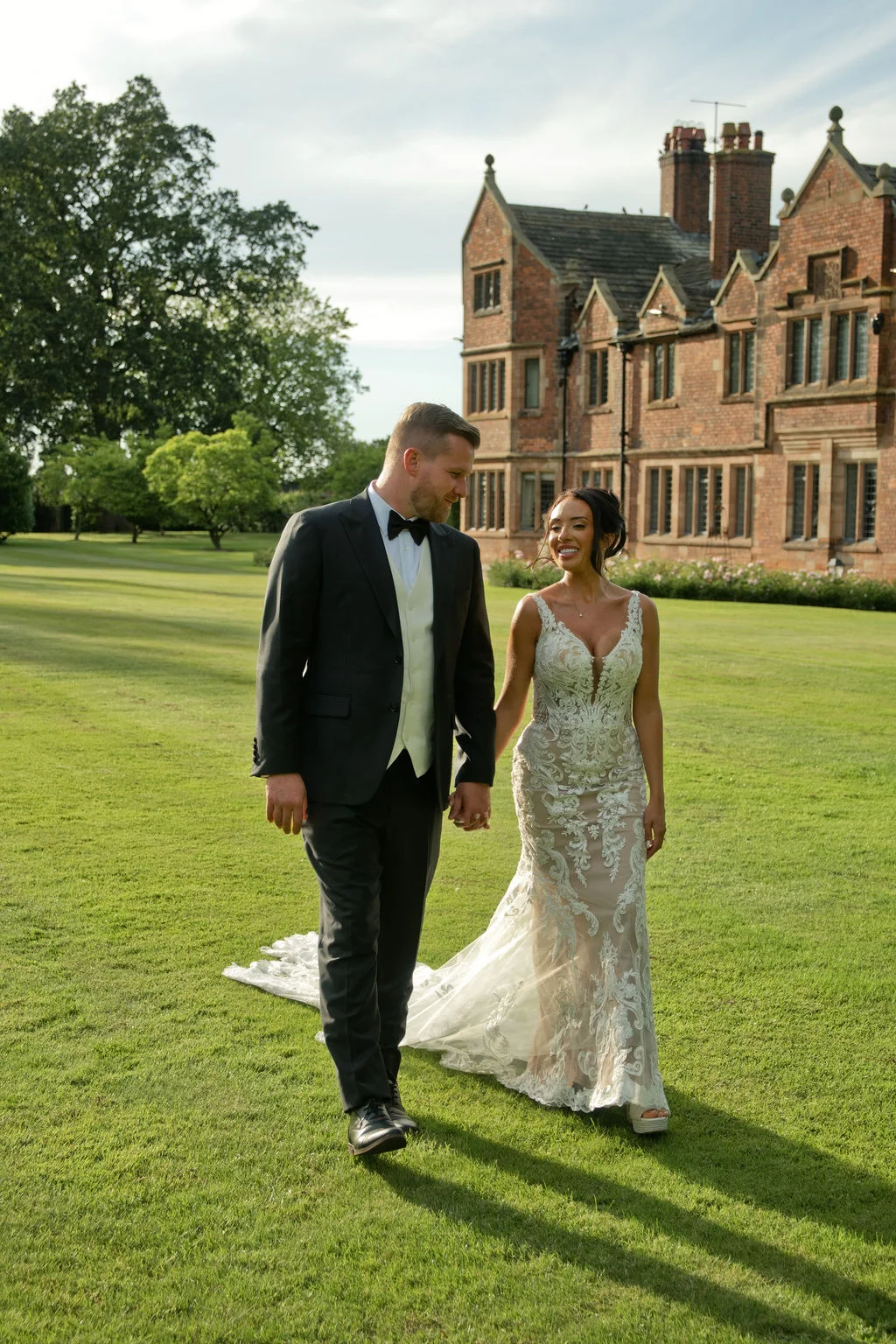 A bride and groom walk hand in hand across a manicured lawn in front of a grand country house during golden hour sunshine.