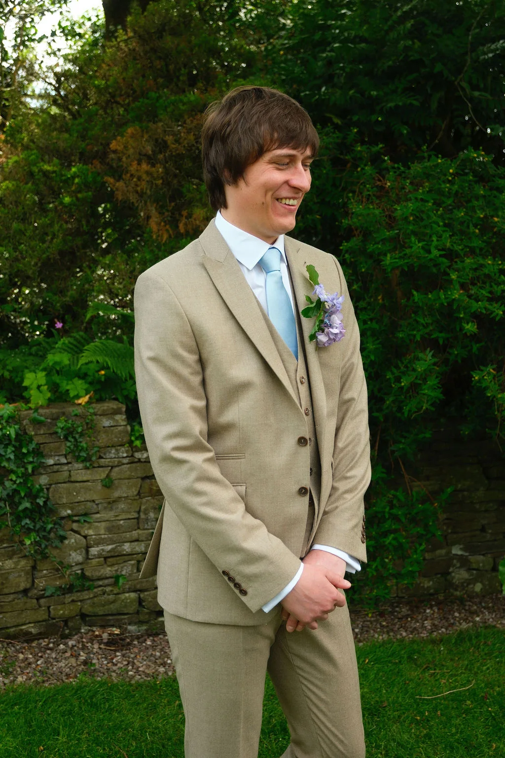 A smiling groom wearing a beige three-piece suit and pale blue tie stands outdoors in front of a dry stone wall.