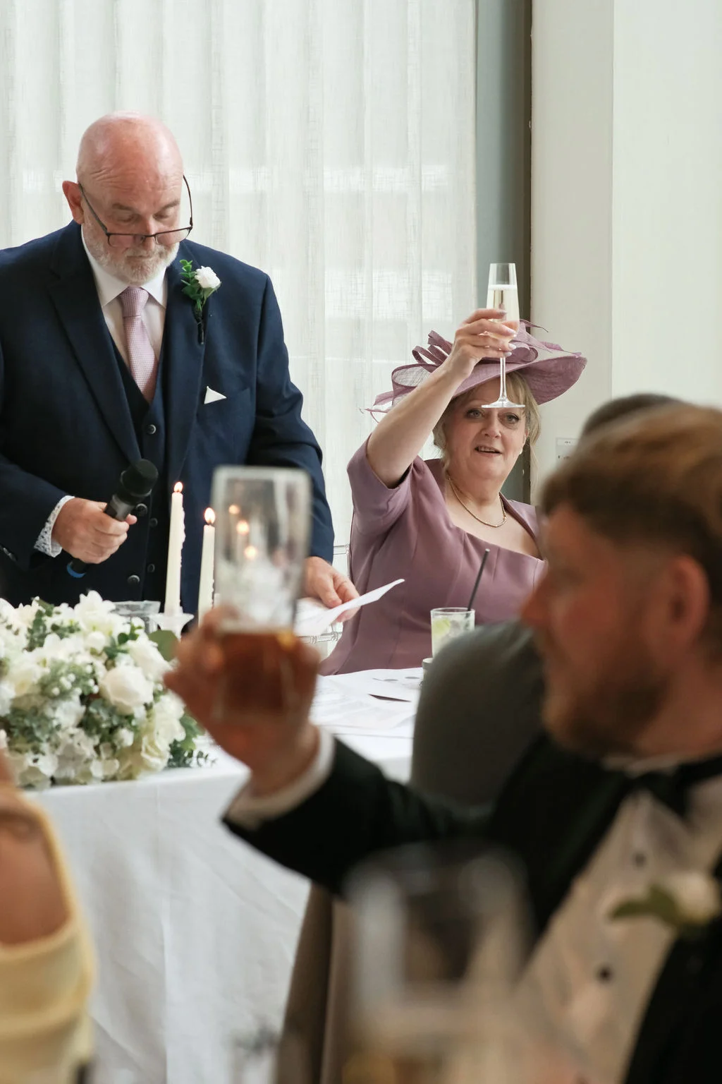 Father of the bride delivers a speech at the top table while mother of the bride raises a champagne flute to toast.