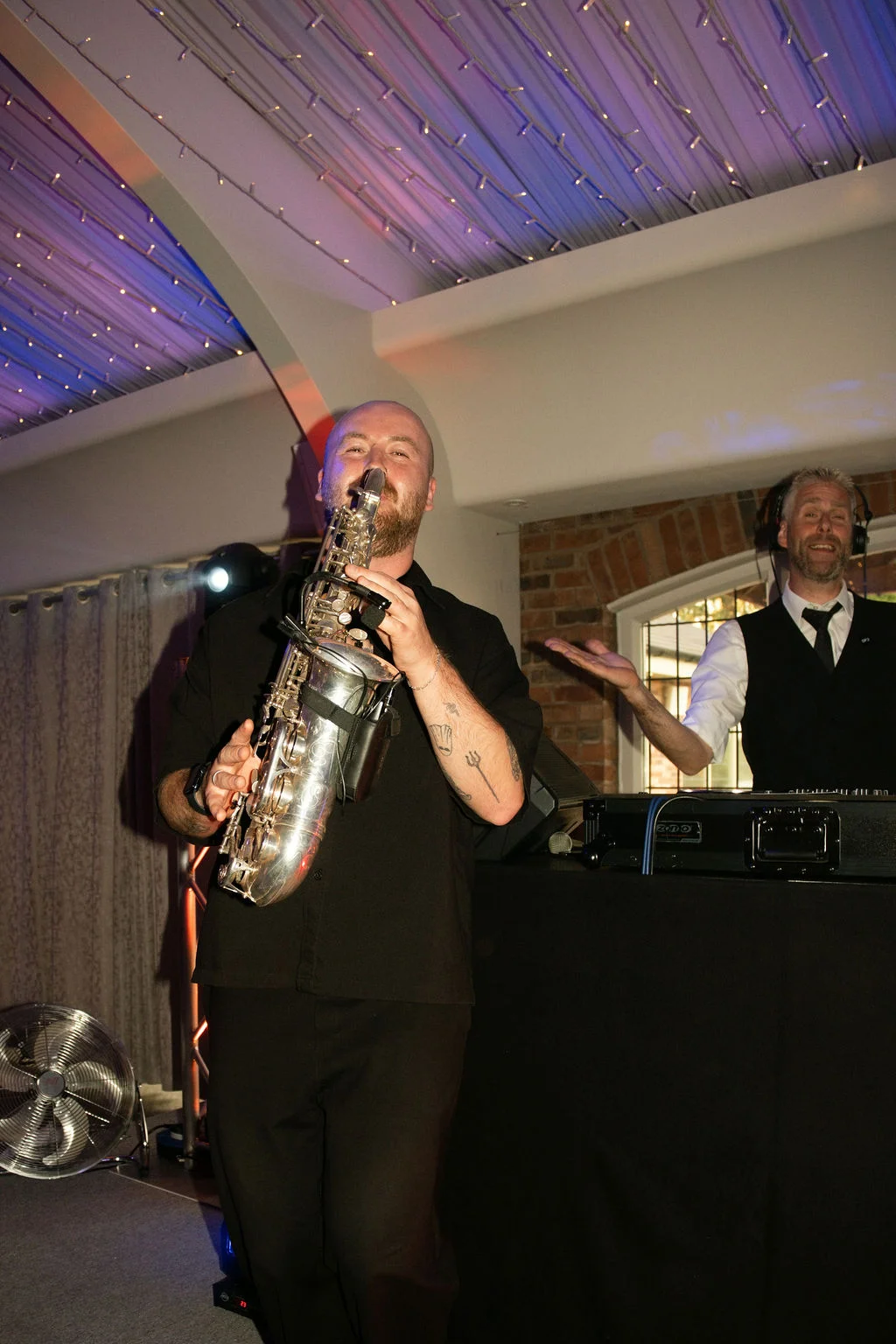 A talented saxophonist plays enthusiastically on the dance floor next to a smiling DJ during a lively evening wedding reception.