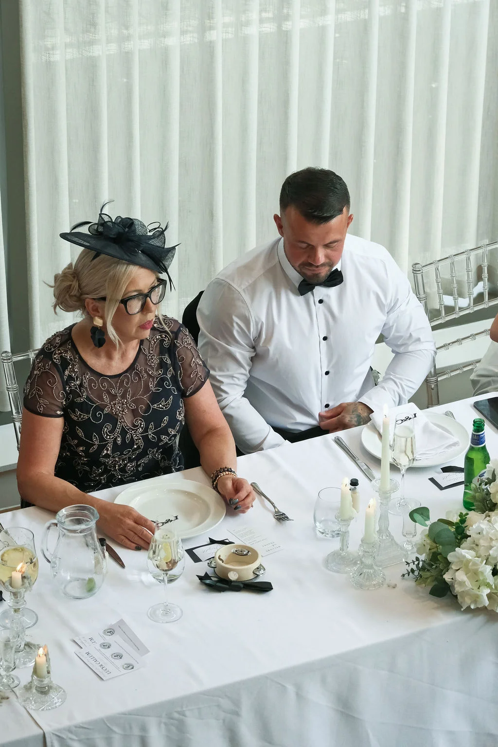 A mother of the bride in a black patterned dress and fascinator sits alongside a groom in a white shirt and bow tie at a neatly set wedding breakfast table.