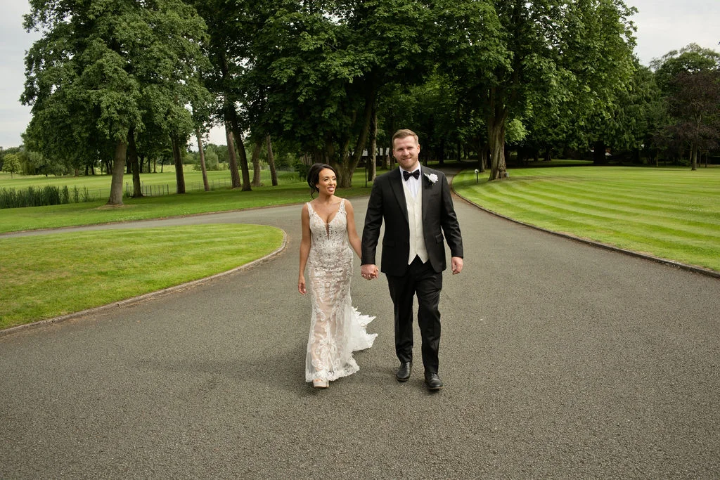 A newlywed couple walks hand in hand along a paved driveway flanked by lush green lawns and tall trees on their wedding day.
