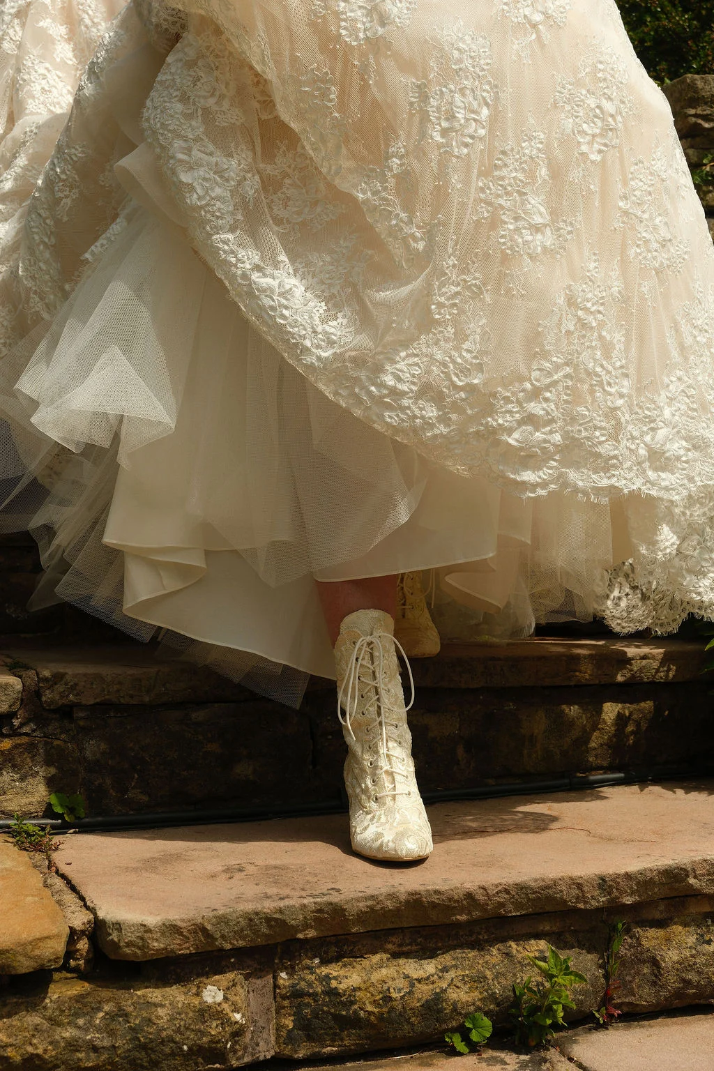 A close-up view shows a bride descending stone steps while wearing vintage-style lace-up bridal boots beneath an elaborate tiered lace gown.