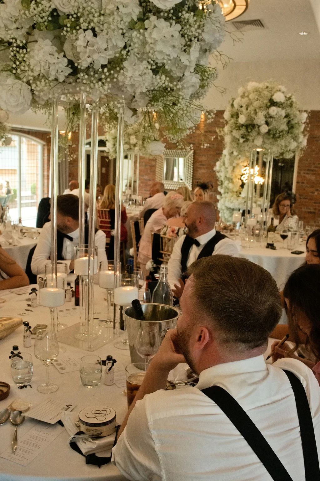 Guests enjoy the wedding breakfast at round tables decorated with tall acrylic stands holding white floral arrangements and glowing candles inside a venue with exposed brick walls.