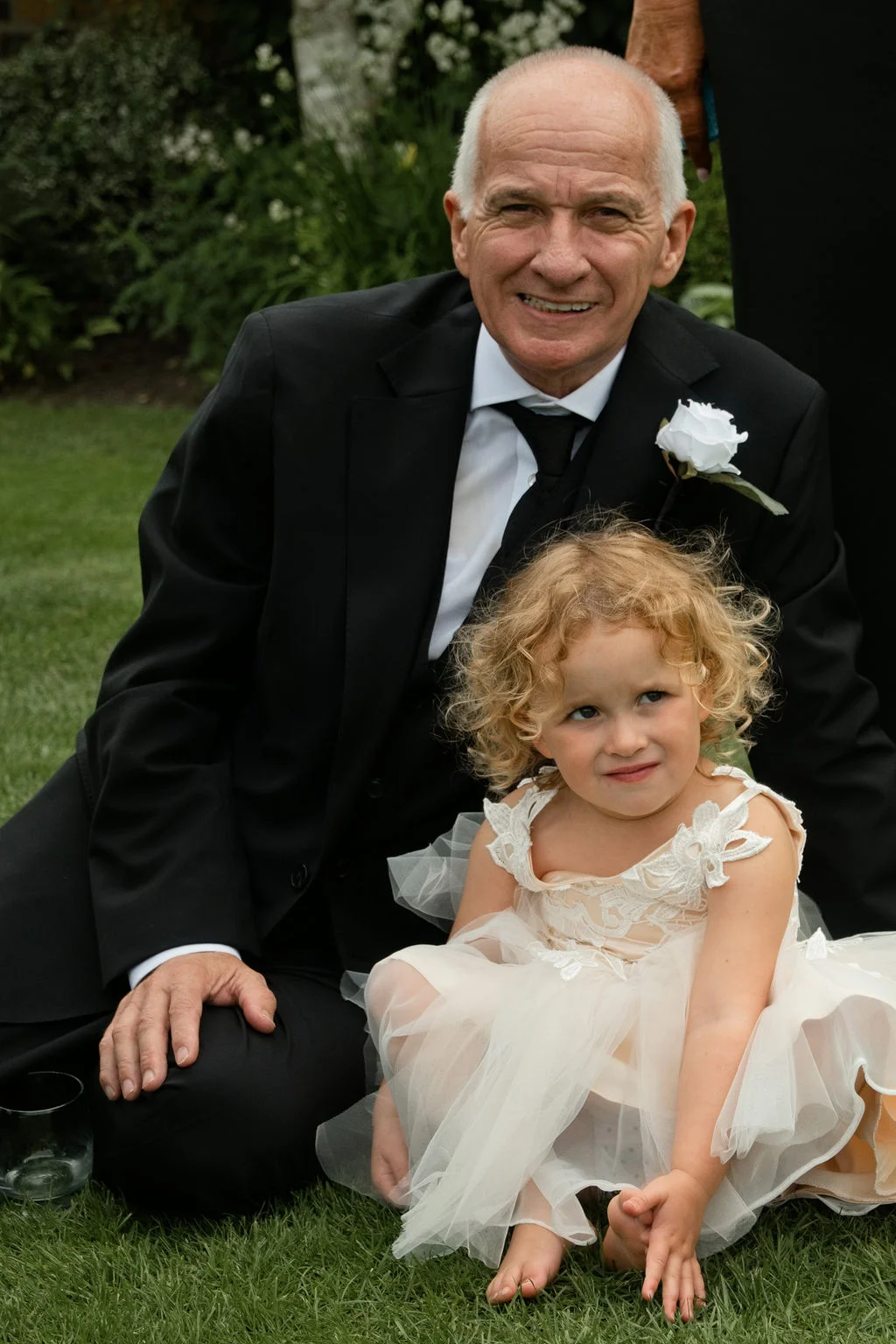 An older man in a black tuxedo and a flower girl in a tulle dress sit together on the grass at an outdoor wedding.