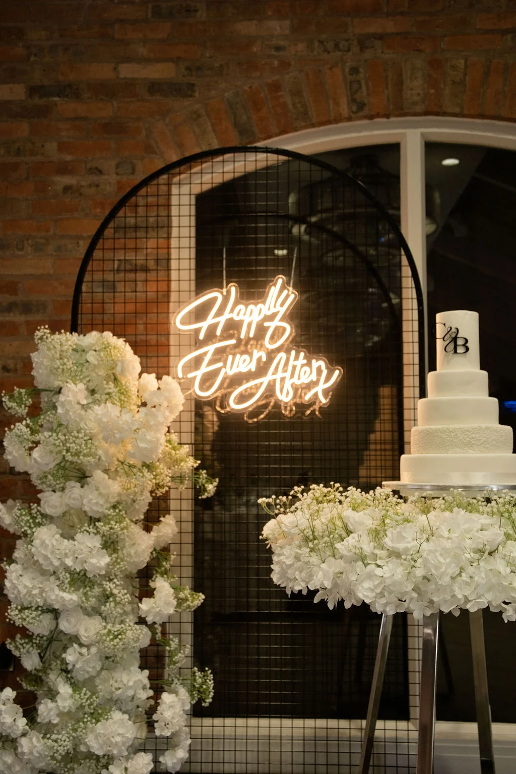 A multi-tier white wedding cake rests on a floral-trimmed stand next to a black grid backdrop featuring a glowing neon sign that reads happily ever after.