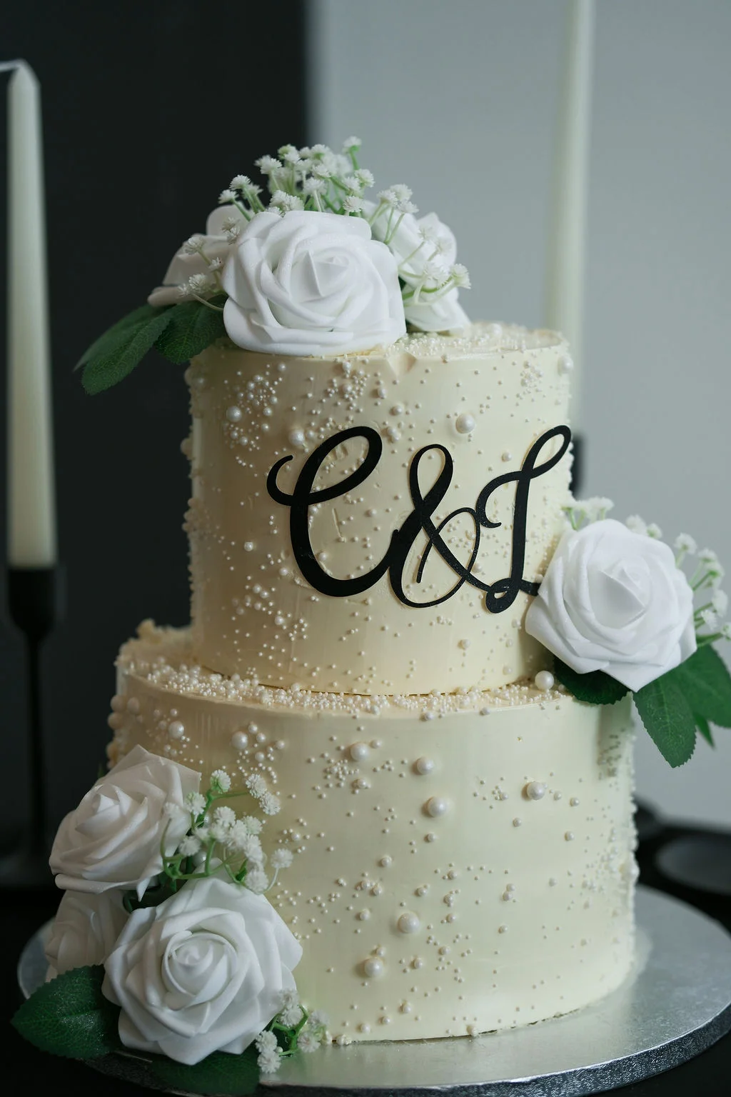 A stunning two-tiered wedding cake adorned with delicate pearl detailing, white roses, baby's breath, and a striking black monogram topper.