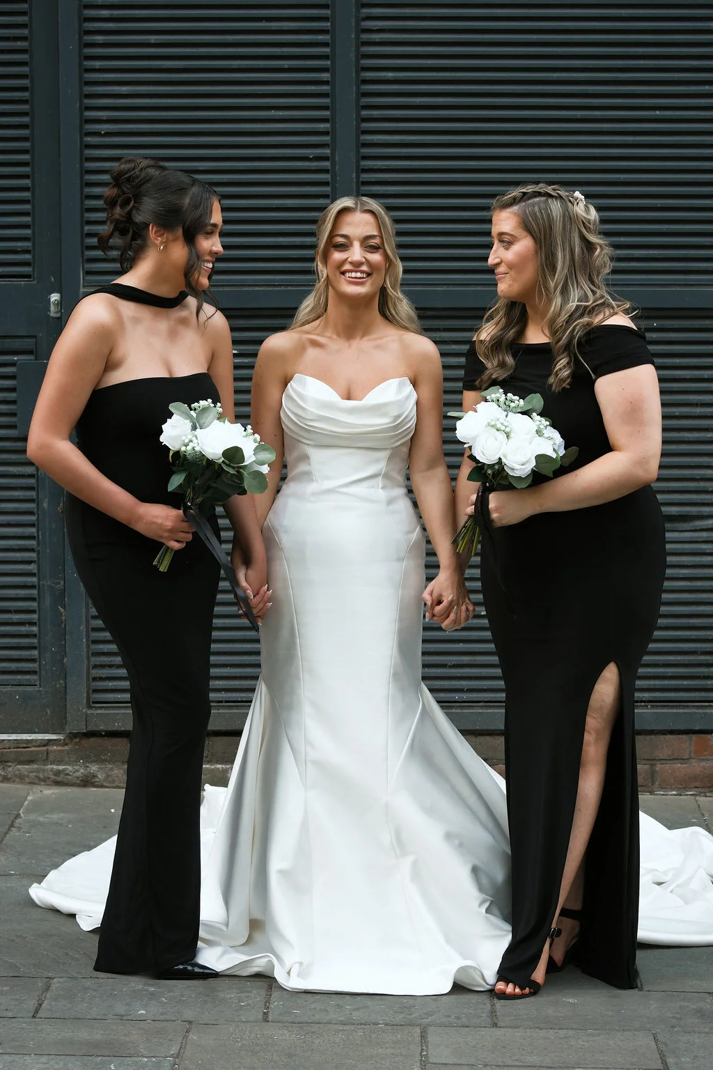A smiling bride in a white satin strapless gown stands in the centre, holding hands with two bridesmaids wearing elegant black dresses and holding white rose bouquets.