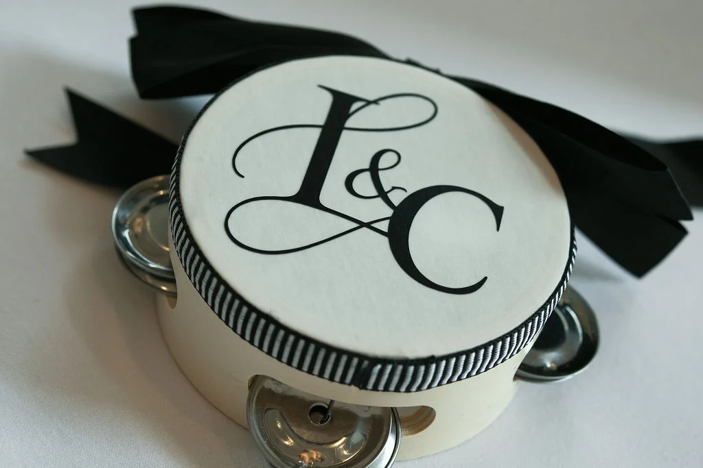 A close-up shot of a custom white tambourine featuring a black monogram and a large black ribbon bow.