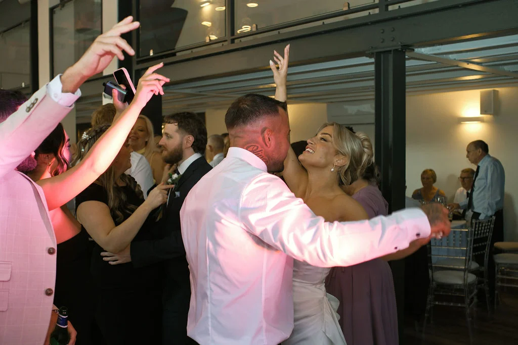 A bride and groom share a joyous first dance on a lively dance floor surrounded by cheering guests under glowing pink lighting.