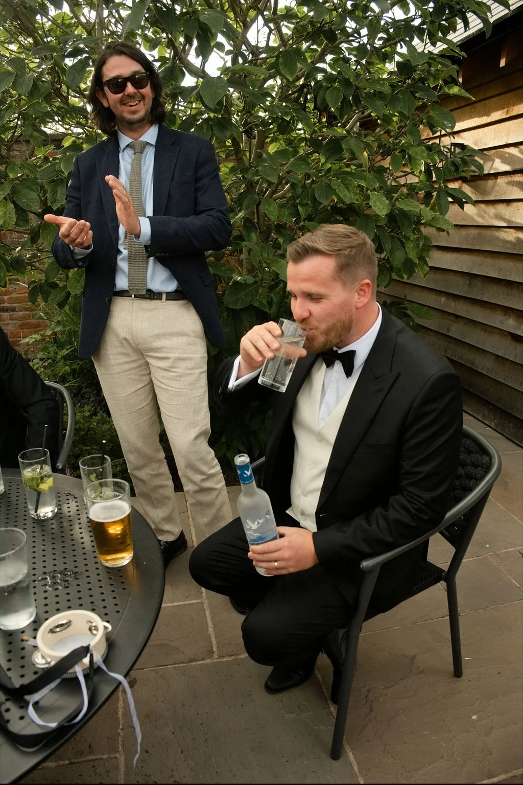 A groom sits at an outdoor table drinking from a glass while a guest in sunglasses and a blazer applauds nearby.