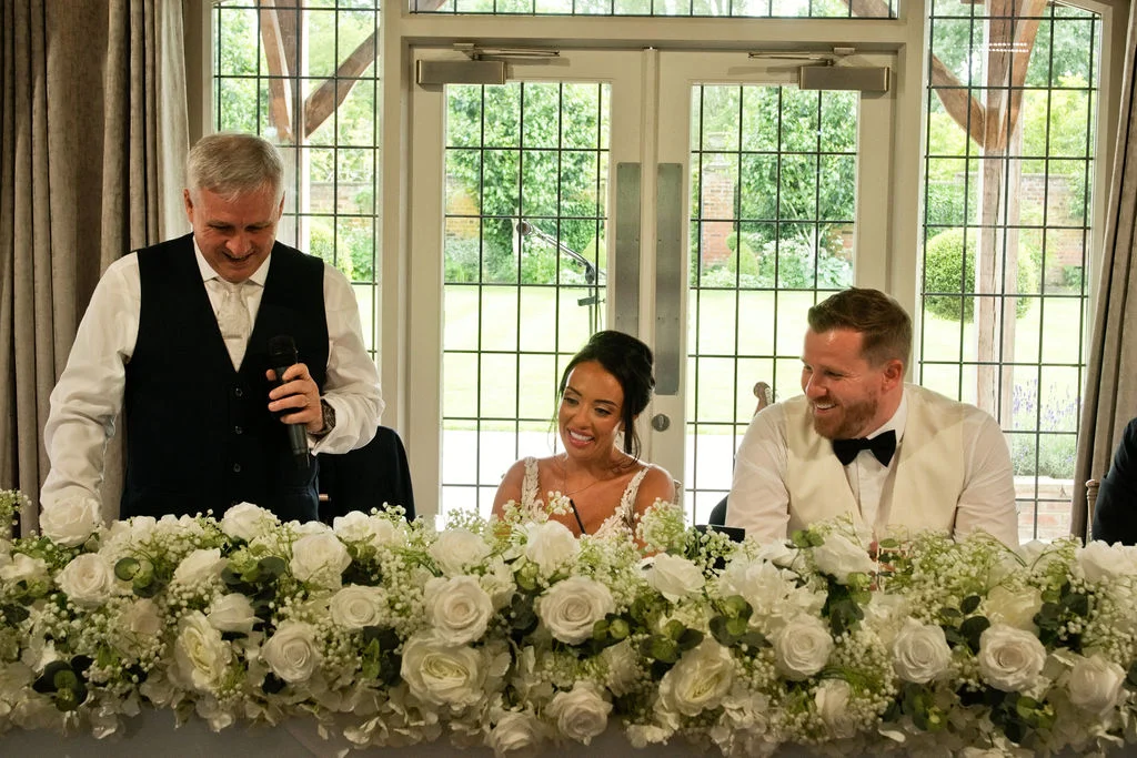 A guest delivers a speech with a microphone at the top table while the smiling bride and groom look on, seated behind an abundant floral arrangement of white roses and baby's breath.
